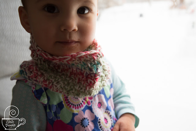 Image of a toddler wearing a multi-colored cowl over a floral sundress and minty long-sleeve oneside. It is snowing outside. The toddler is next to the closed glass door.
