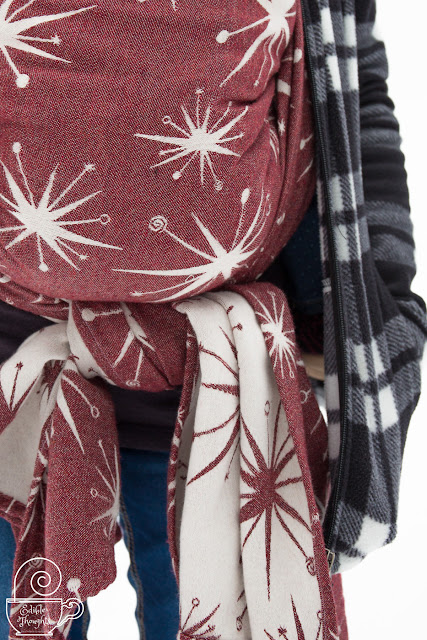 Image of a maroon and ecru retro star patterned woven wrap of the back of a sleeping toddler in the wrap with the image focused on the knot under toddler's bum. On the side is the Mama's black and white fleece with hand in pocket and white snow in the background.