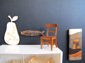 Modern dolls' house miniature display wall in a gallery. On display are a wooden pear, fish and chair, and wall plaque.