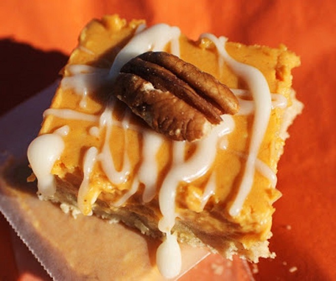 these are an easy how to make pumpkin cheesecake bars. These are creamy filled bars with a cookie crust on the bottom. Drizzled with powdered sugar frosting and pecans on top. They are on orange napkins and a Thanksgiving and Halloween Fall Treat.