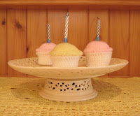 A cream, filigree-patterned, porcelain cake stand rests on a yellow filet crocheted mat. On top are three crocheted cupcakes with white bases and tops of yellow and pink. Each cupcake holds a birthday candle. The pine wood in the background is the cupboard on which the cake stand is displayed.