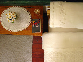 View from above of a modern miniature doll's house bed with white cotton embroidered bedding . On the bedside table is a glass of water, a diary and reading glasses and a vase of daisies.