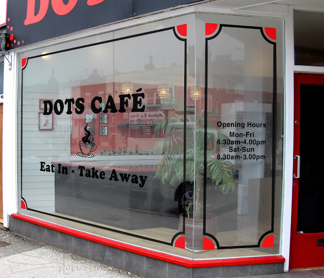 Black vinyl boarder around two display windows with red quartered circles in the corners. Bold black text with 'Dots Café' & a steaming coffee logo in the main window. Shop opening times in vinyl on the second window that can be changed easily.