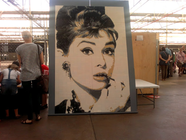 A giant portrait of Audrey Hepburn made of lego bricks.  It is taller than a person.