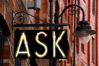 A sign hanging from a lamp attached to the side of a brick building. It is black and reads 'ASK' in white and gold capital letters.
