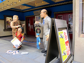 Three figures in front of a modern dolls' house miniature cinema lobby. One man is attacking an usherette, the other is holding a marionette.