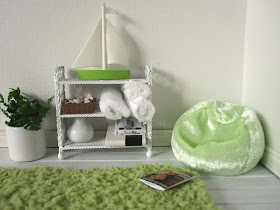 Corner of a one-twelfth scale modern miniature holiday house with a green fluffy rug, bean bag and a shelf holding towels and books.