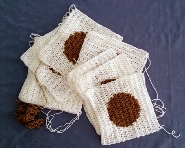 A stack of 7 squares crocheted in rows (not granny squares which are crocheted in rounds). The squares are white with a big tan circle in the middle, using intarsia technique. The stack of squares has been spread out across a blue background. The squares are overlapping. The loose ends can be seen because they have not been woven in yet.