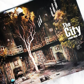 Front cover of the book 'In the city' by Lori Nix showing a two-storied library space in a state of neglect, with a tree growing out of the floor and through a hole in the ceiling.