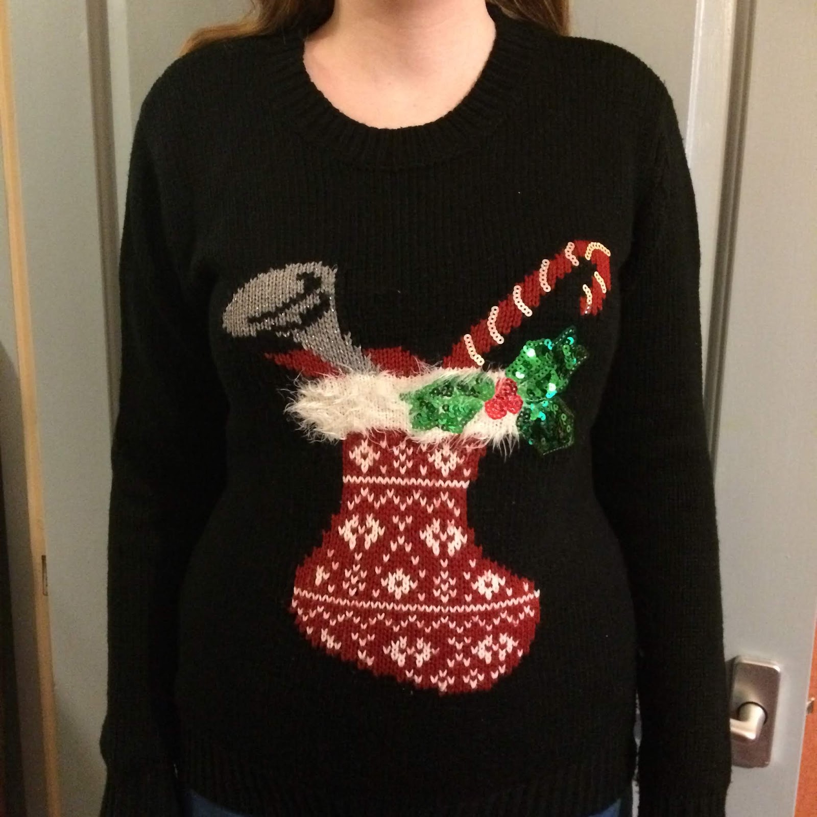 A black jumper featuring a red stocking with a white nordic pattern, with a white fluffy trim, and sequined holly. There's a candy cane and a horn.