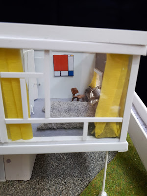 View through the window of a 1/48-scale mid-century modern house, showing a bedroom in white with accents of bright colour. Underneath are patches of concrete, gravel and grass and to the right is a ramp leading upwards from the garden.
