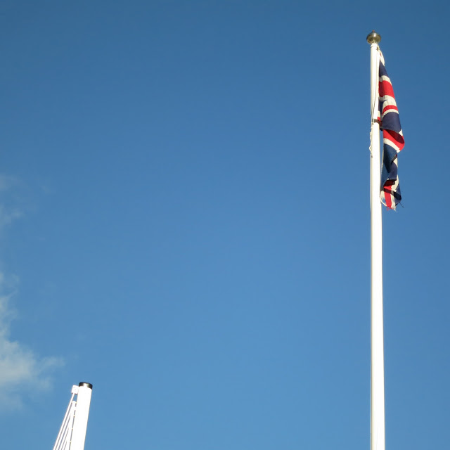 Top of raised barrier and part of top of flag pole with Union Jack against a blue sky.