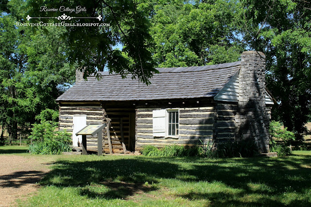 Photo of an old slave cabin at the Sam Davis Home in Tennessee.  There is what is called a "dog trot" or opening down the hallway separating the cabin into two pieces with a roof covering all. The part on the left has a white door and the right has a small window with a single white shutter. There s a stone fireplace on the right and left of the cabin.  by RosevineCottageGirls.com