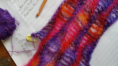 silk mohair scarf in progress with a bamboo crochet hook resting next to it on top of some handwritten pattern notes. At the left of the photo is a ball of purple silk mohair yarn.
