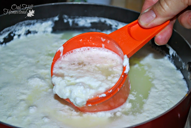 A woman's hand using an orange measuring cup to gently scoop the curds out of a large pot.