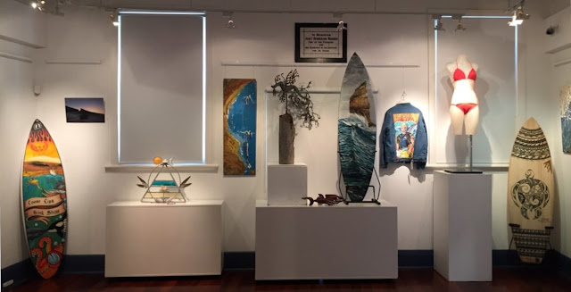 A selection of artworks (left to right): painted surfboard, surf photograph, mixed media sculpture, painting, metal sculpture, woodworked pieces, painted surfboard, embellished denim jacket, crocheted bikini, painted surfboard, painting (part).  On the wall in the background above and centre is a foundation stone or plaque of the building. The writing is blurred.