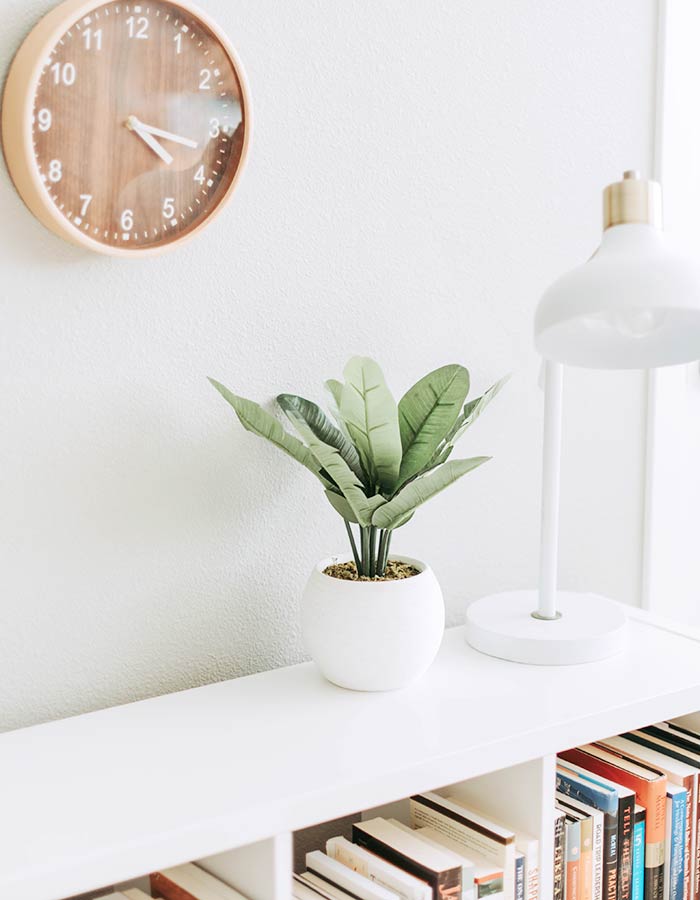 Planta ornamental artificial na decoração da estante de livros; ficou um charme com a abajour moderna e o relógio de madeira na parede.