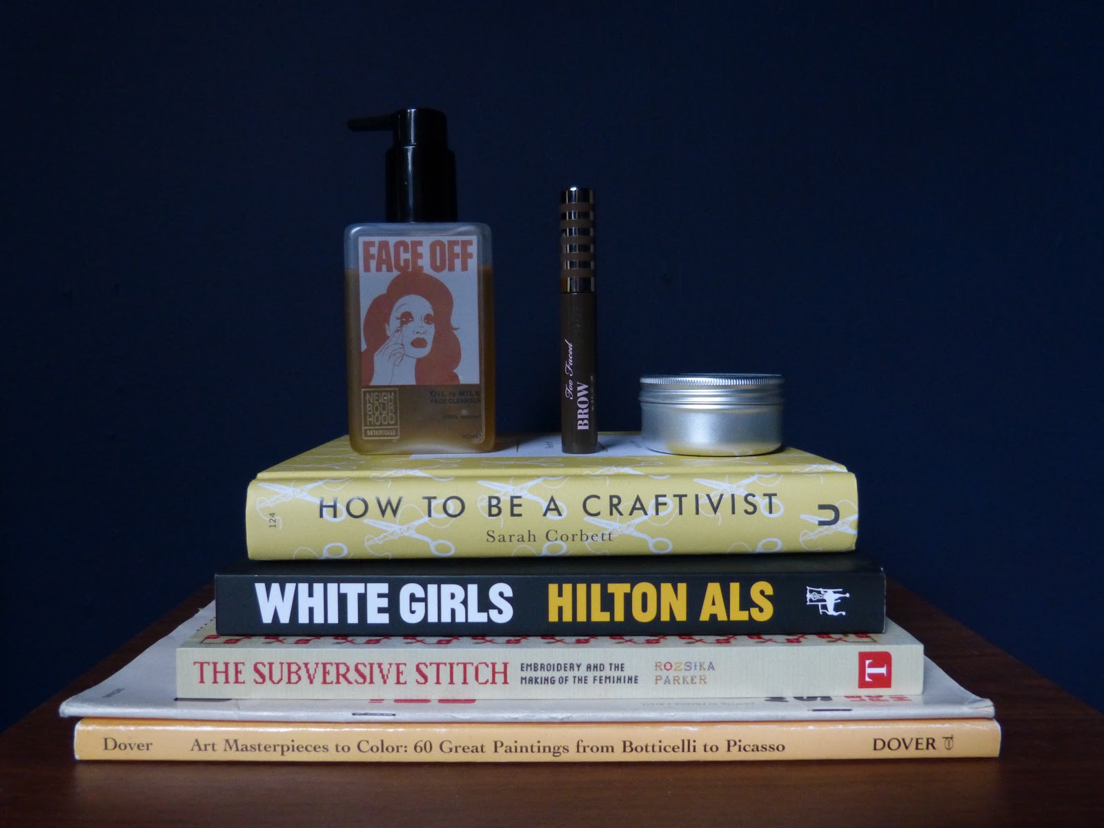 The picture shows a pile of books (from below to top: a colour book, a vintage magazine, The Subversive Stitch by Rozsika Parker, White Girls by Hilton Als and How To Be A Craftivist by Sarah Corbett. On top of the pile of books stands various makeup products. From left to right: Face Off Oil to Milk Cleanser by Neighbourhood Botanicals, Brow-Quickie Brush On Fibre Gel by Too Faced and Ultrabalm by Lush. The background is blue.