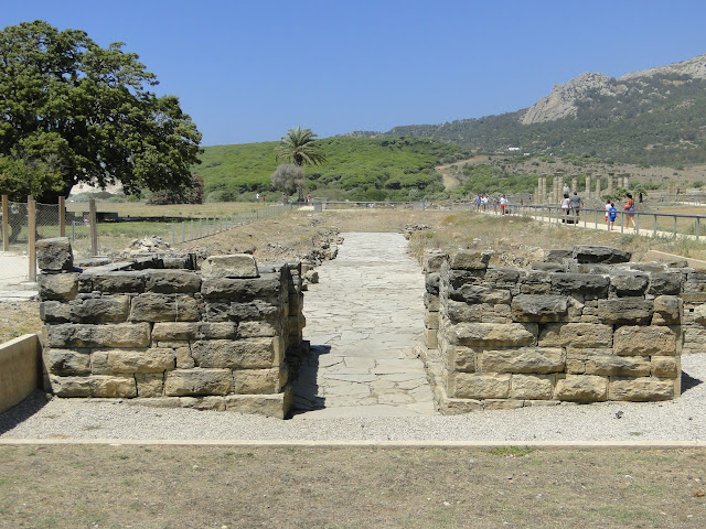Portada de entrada de piedra de ciudad romana en ruinas, con vegetación mediterránea y una montaña al fondo.
