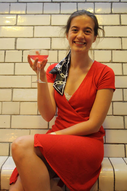 Abbey sits on a white tiled wall, wearing a red dress and matching shoes. She smiles and holds up a Cosmopolitan cocktail