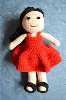 The AKA Amigurumi project. A little doll with straight black hair, black button eyes with eyelashes, a red dress and black sandals.