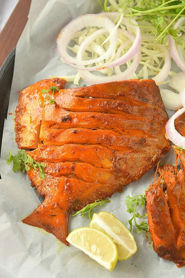 A  masala fried fish served on white paper with lemon wedges and onion rings
