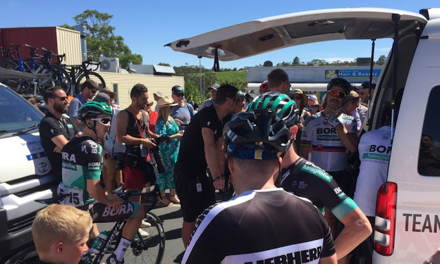 Behind the team van, riders, technicians and onlookers are milling around. Peter Sagan is underneath the shade of the tailgate adjusting his helmet and radio earpiece equipment..