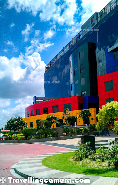 Adobe India has one of the most colorful building in Noida city near to Capital city of India. This Photo Journey shares some of the mobile clicks of Adobe Noida campus from outside. Let's check out and know more about the same.Adobe Noida building with these vibrant colors was first building in sector 25A of noida. Till date, there are only two buildings in the city center sector of Noida. After one of the biggest real estate deals in Noida, Wave group has acquired rest of the sector now and building various facilities around the same. Here is a photograph of beautiful landscapes in the campus - Water fountains, green plants, colorful tiled platforms etcWhole building remains clean most of the times because of continuous cleaning efforts in campus. This building is also one of the rare green buildings in the city. Adobe Noida building has been awarded various recognitions for green efforts in the city. Adobe office in Noida is just in front of Sport-Stadium in sector 21A and Reliance building in sector 24. It's on the corner of sector 25A, facing Sector 24 on one side and sector 21A on the other side.Campus has small golf ground, half basketball court, Volleyball court & lawn tennis facilities apart from various recreational options inside the building. 
