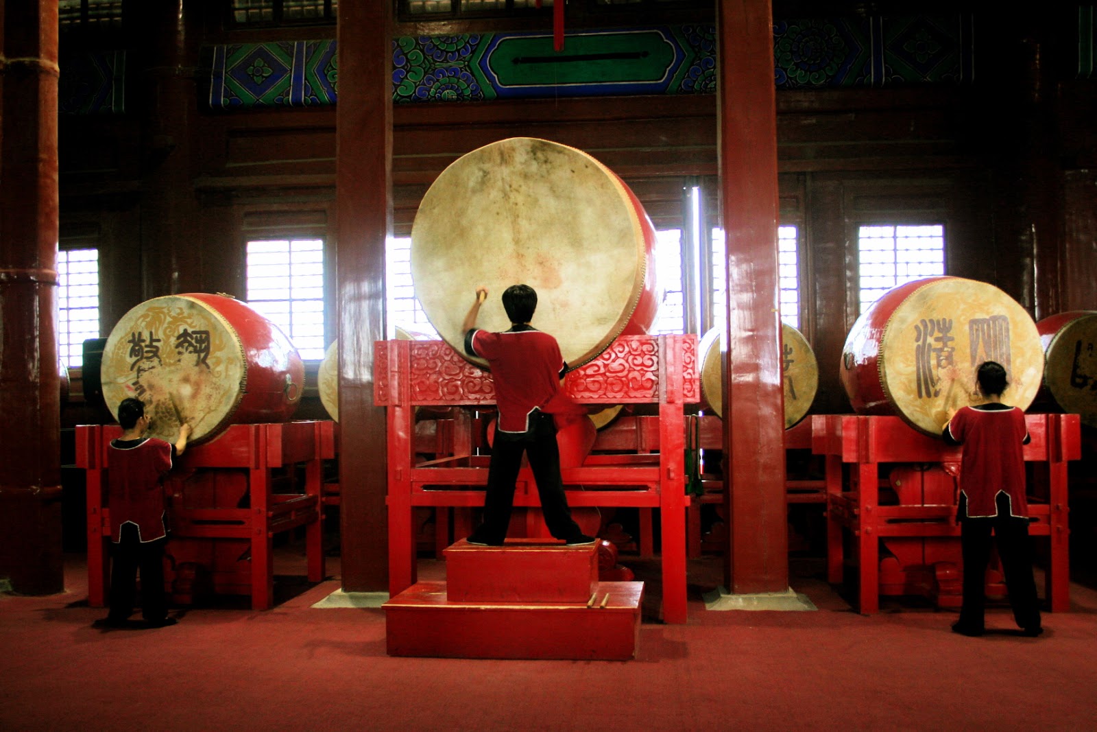 Tambores chineses, em um ambiente em tons vermelhos, tocados por três pessoas ilustra este post sobre o Shijing, o Livro das Canções.