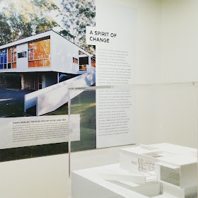 Architect's model of a modernist building in a perspex case in front of a large photo and information about it on the wall behind.