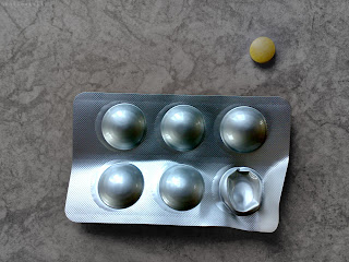 Front side of silver coloured blister pack containing six 85mg Potassium Iodate tablets in blister pack. One tablet has been removed from pack showing it's smooth side. The tablet has begun to yellow with age.
