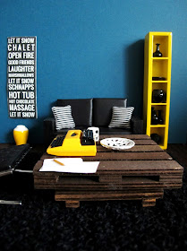 Modern one-twelfth scale miniature lounge in teal, yellow and black, with a typewriter sitting on a pallet coffee table.