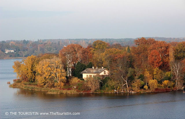A white building on an island surrounded closely by trees with vividly coloured leaves; autumn light.