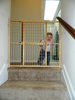 A child's gate at the top of the stairs with a tot standing behind it