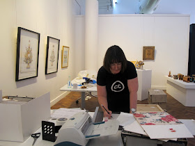 Woman in a gallery during install, marking something with a pencil on  a table in the middle of the gallery.