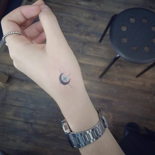 Um preto lua crescente é apoiado por uma estrela azul no presente simples, tatuagem com tinta para o usuário com a mão esquerda.
