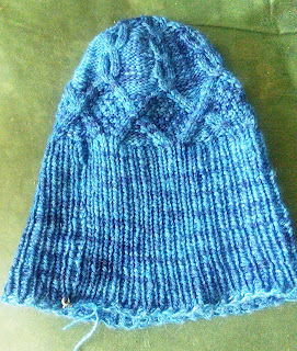 A blue hat laying flat.  The hat is knit in worsted-weight yarn, and cabled.  It has a long bottom edge knit in 1 by 1 ribbing. 