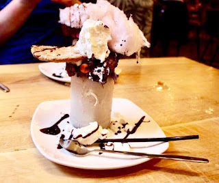 A tall milkshake style clear glass that is rimmed at the top with dark brown Nutella, light brown chocolate flake and colourful smarties in different colours,  filled with a scoop of dark brown chocolate ice cream blended with white vanilla milkshake to give a light brown colour, topped with a wedge of dark brown chocolate fudge cake, light brown chocolate chip cookie, white squirty cream, light pink candy floss all on a white circular plate with squiggles of dark brown chocolate sauce on a wooden table on a light background. 