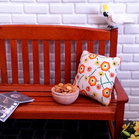 Modern dolls house miniature scene of a garden bench with cushion, a bowl of chips and an IKEA catalogue. On the back of the bench a cockatoo eyes up the chips.