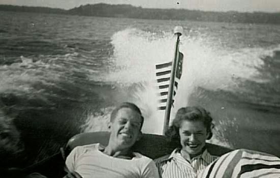 Black and white photo of young man and young woman seated next to each other in the back seat of a running motorboat