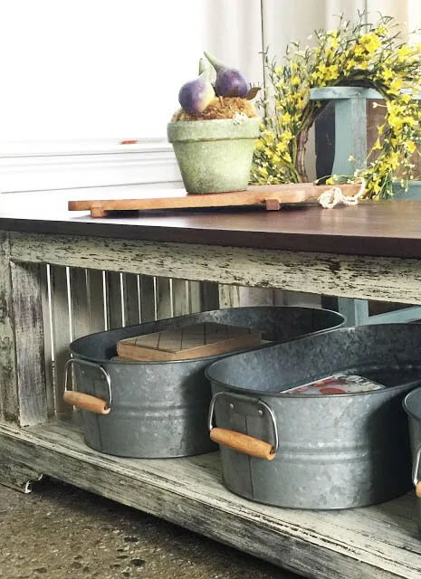 Two galvanized tubs with handles tucked under a table with books and magazines inside