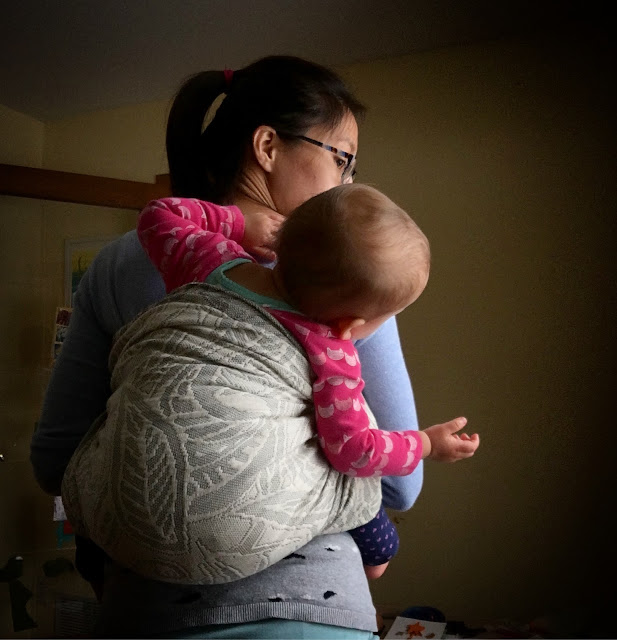 [Image from back of a tan skin bespectacled Asian woman wearing a toddler on her back using a gray botanical patterned woven wrap carrier in a no-shoulder carry. Toddler is leaning to the side to tell Mama something. Mama is listening.]