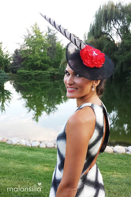 Invitada boda con vestido estampado geométrico en blanco y negro, tocado tipo plato en negro y flor rojo con plumas de faisán posando con escote en la espalda y un lago detrás