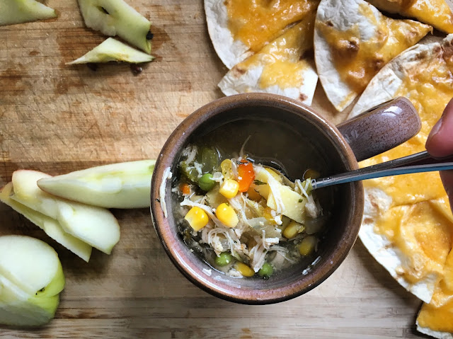 [Image from above of a tan ceramic mug filled with hot chicken noodle soup full of veggies and a silver spoon. It’s sitting next to sliced apples and melted cheddar on baked flour tortillas. They’re on a wood cutting board.]