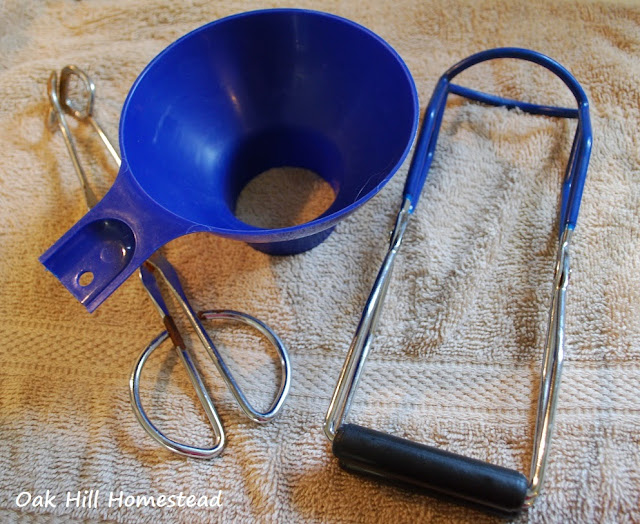 Canning funnel, jar lifter and metal tongs on a beige kitchen towel.