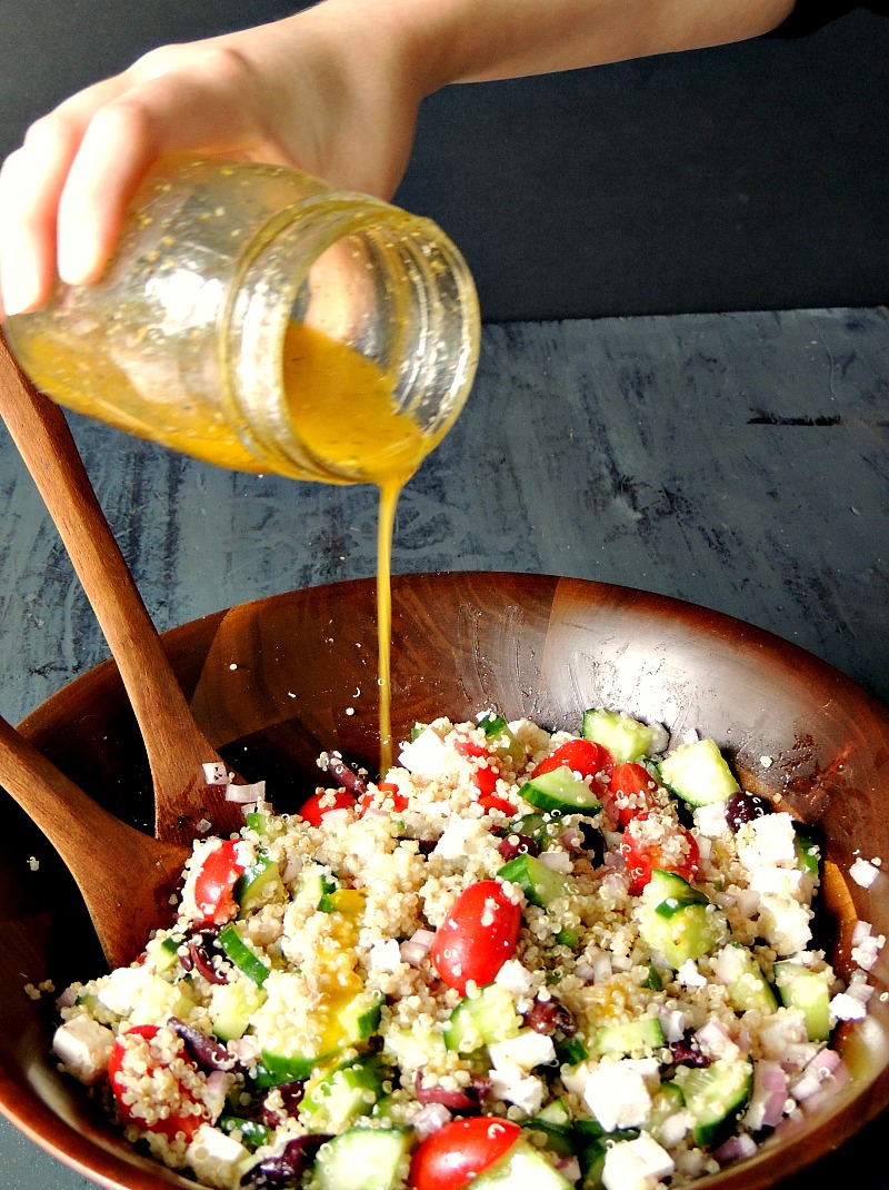 Pouring the dressing into Mediterranean Quinoa Salad in a wooden salad bowl on a blue table.