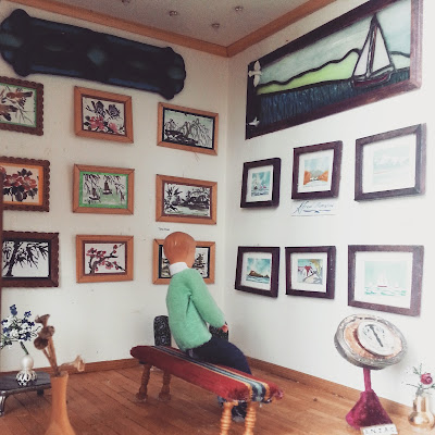 Miniature art gallery, with various pictures displayed on the walls. There is a bench in the middle, with a doll sitting on it looking at the art.