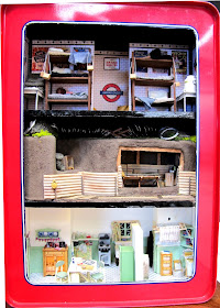 Rectangular biscuit tin with the top on the side, cut out to show an air-raid shelter in a tube station, an anderson shelter and a 1940s kitchen scene.