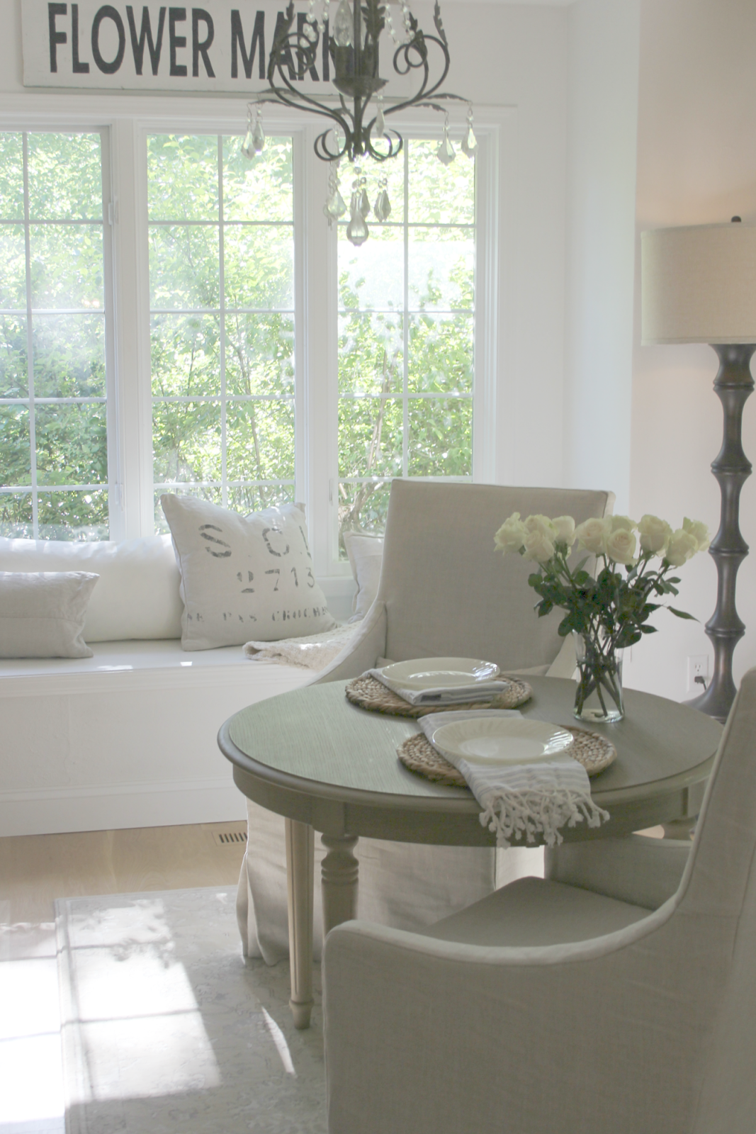 Serene breakfast room with window seat, round dining table, Belgian linen slipcovered arm chairs, and vintage Flower Market sign. #hellolovelystudio #kitchendecor #breakfastroom #windowseat #belgianlinen #vintagestyle #modernfarmhouse #serene #neutraldecor