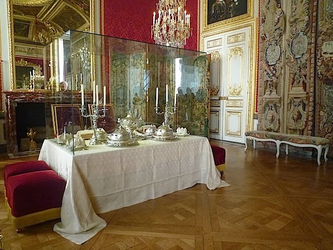 Photograph of a restored salon at Versailles. The decor is extravagant with a chandelier and ornaments on the walls and doors. There is a dining table with silverware and china. Around the table are two chairs and four stools.